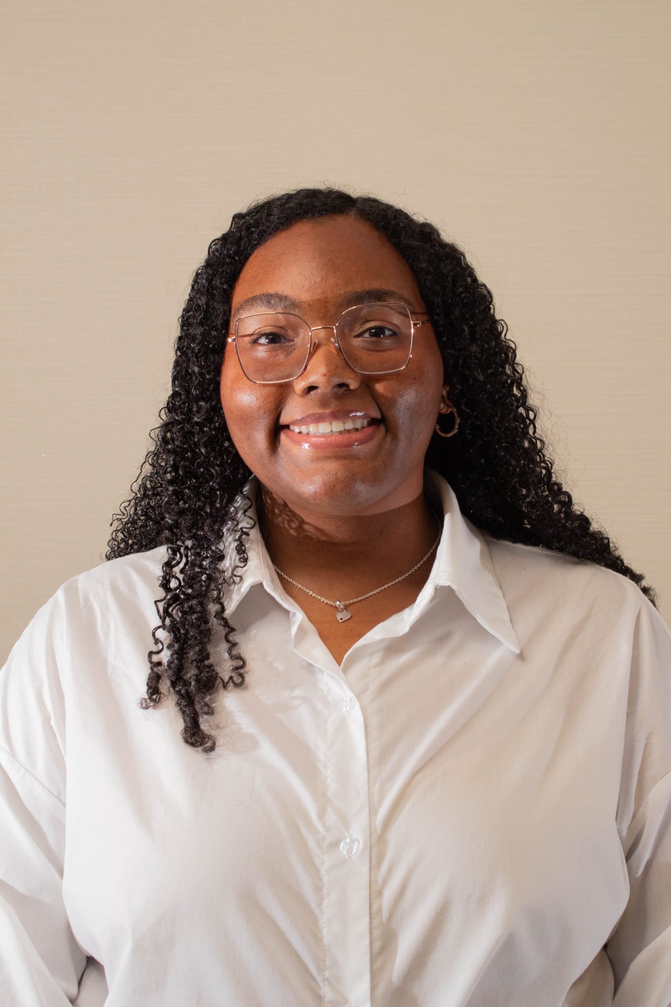 Amaris smiles at the camera. She has dark skin and long black hair that hangs in tight curls past her shoulders. She is wearing thin-framed square glasses, hoop earings, and a silver pendant necklace. Her shirt is white and buttoned-up and collared. She is standing in front of a white background.