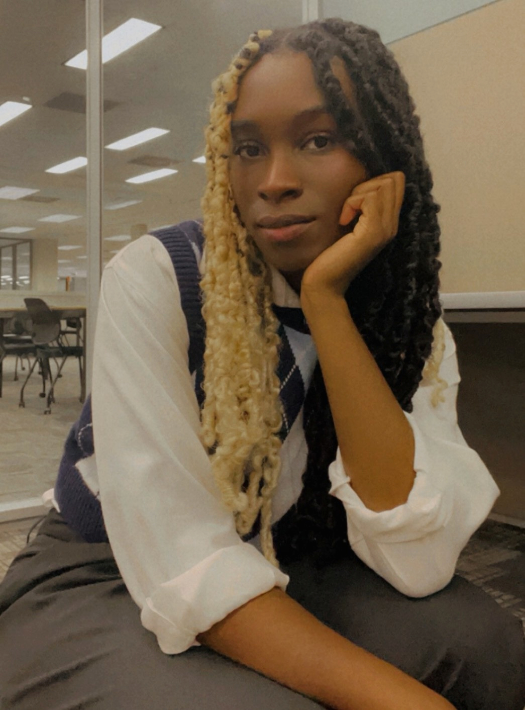 Angelique sits looking at the camera. One elbow rests on her knee as her hand props her chin. She is wearing grey slacks and a white collared shirt with a blue argyle sweater vest. Her hair is two different colors, separated down the middle part. One half is bleached blonde and the other is black. In the background, one can see study tables and chairs.