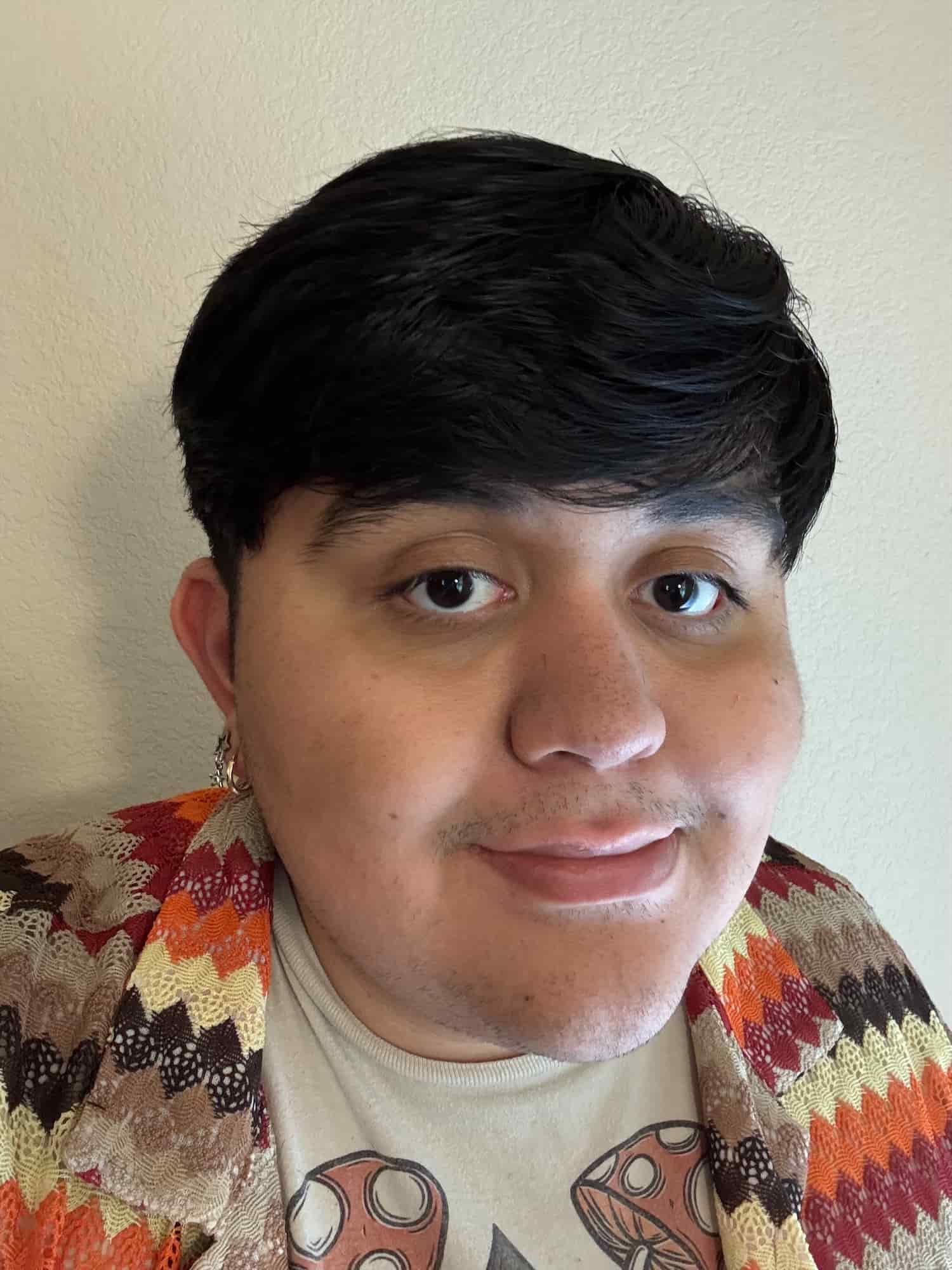 Elijah looks at the camera with a small smile. In one ear, one can see that Elijah has multiple hoop earrings. He is a Hispanic man who has black hair and dark brown eyes.He is wearing a shirt with mushrooms barely visible in the frame and a striped, zigzag patterned, knitted cardigan. The colors in the patterned cardigan are red, orange, cream, black, brown, and grey. In the background is a white wall.