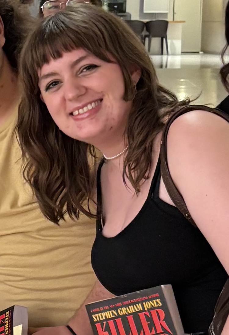 Celeste smiles at the camera; she is wearing a black sleeveless shirt and a pearl choker necklace. She has brown wavy hair with fringe bangs cut straight across her forehead. She is holding a book by Stephen Graham Jones called "Killer." In the background, is a large room with linoleum flooring.