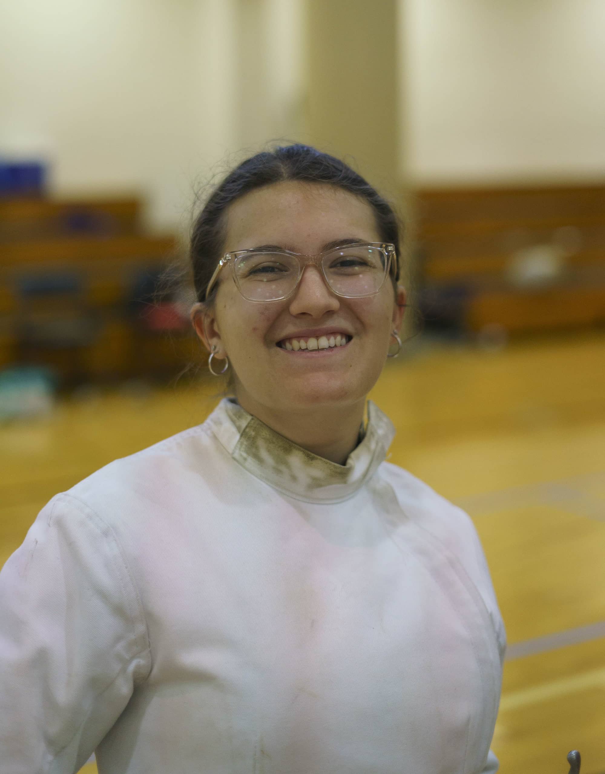 Emma Dore smiles at the camera; she hasdark brown hair pulled into a low ponytail. She is wearing clear-framed glasses and has her ears double-pierced. She is wearing a white fencing uniform, and in the bottom right corner of the photo, one can barely see the handle of her fencing foil. In the blurred background, one can see that she is standing in a gymnasium.
