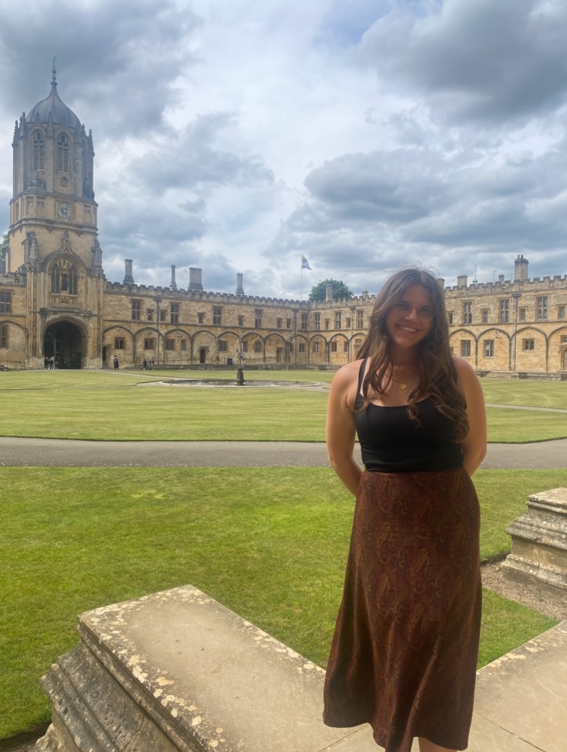 Emma stands on a stone platform in front of a large castle. Between her and the castle is a green lawn with a large, circular fountain. She has long, wavy brown hair and is wearing round-framed glasses. She is wearing a black tank top with thin straps and a long brown skirt. The sky in the background is overcast.