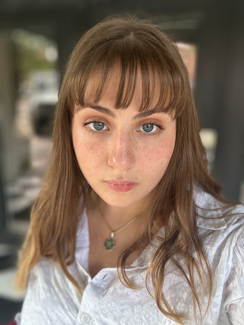 Evelyn stares at the camera. She has blue eyes and light brown, straight hair that hangs past her shoulders. She has bangs that cover her forehead. She is wearing a white collared shirt and a pendant necklace. The background is blurred.