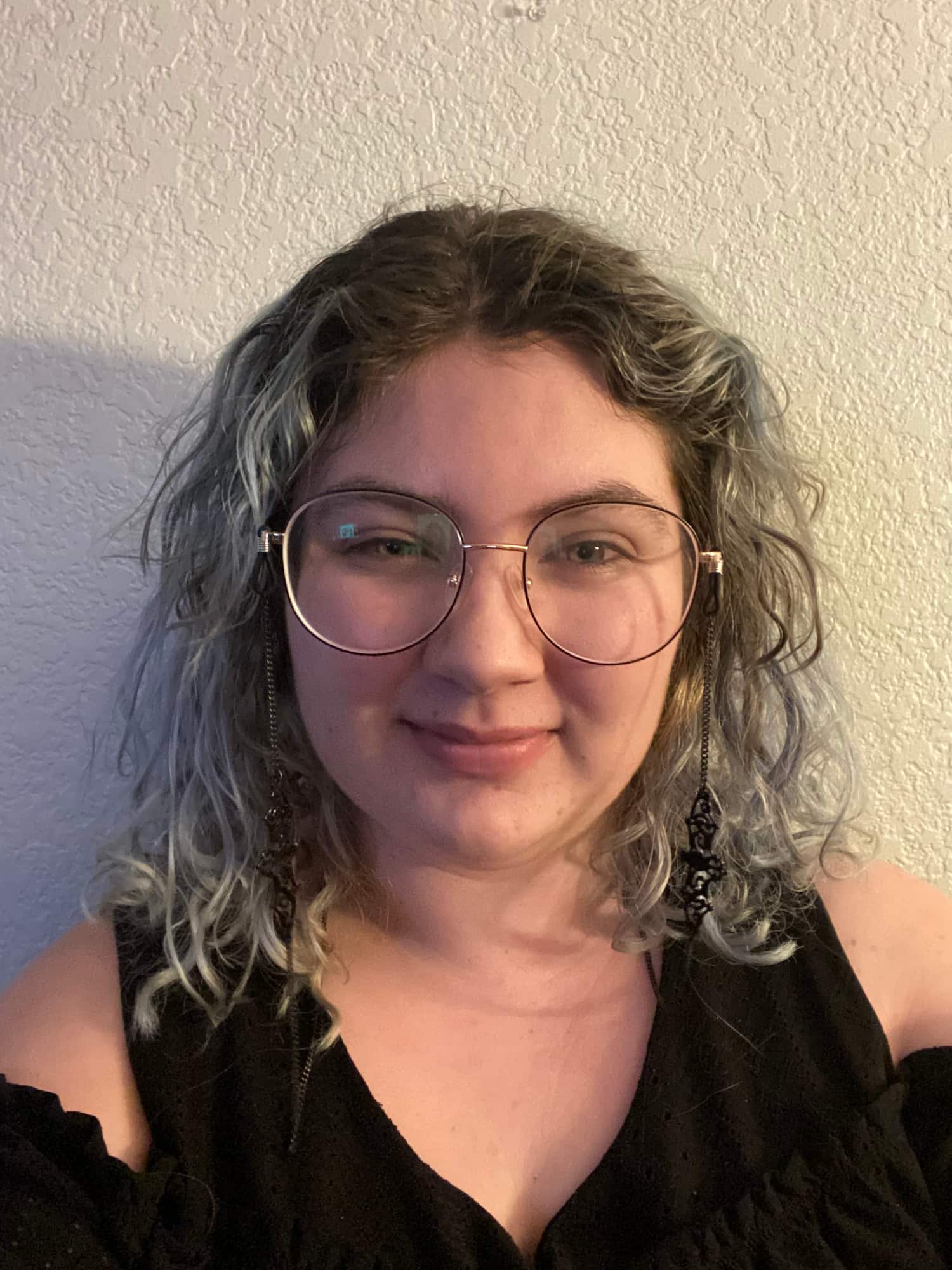 Fable is standing in front of a white wall. They are wearing a black shirt and round, thin-framed glasses. Their glasses have a black, ornate chain hanging from them. Their hair is curly and dyed gray at the ends.
