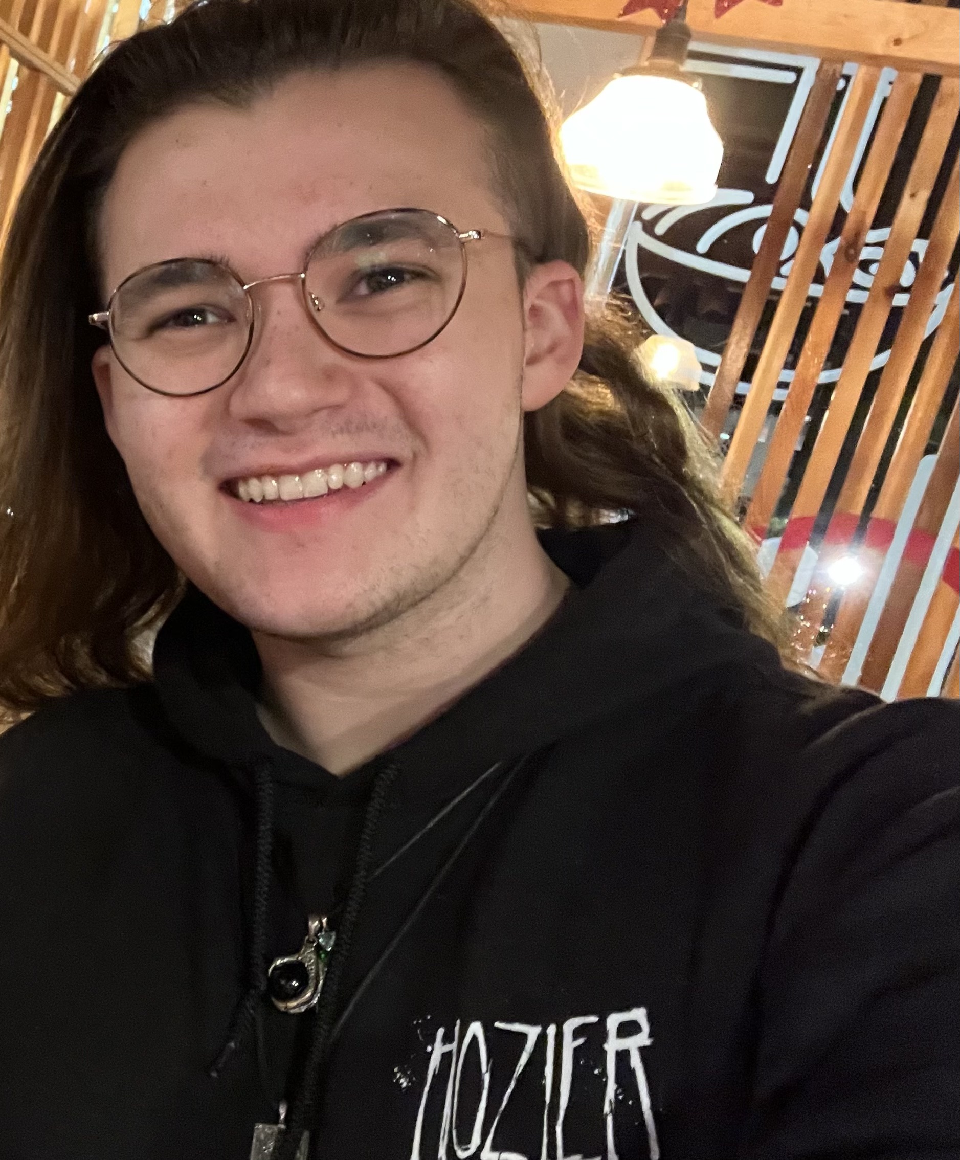 Grant smiles at the camera. He is wearing a black jacket with Hozier written on the left chest. He has circular, thin-framed glasses and a black pendant necklace. He has dark brown eyes and straight, medium-brown colored hair that reached past his shoulder. 