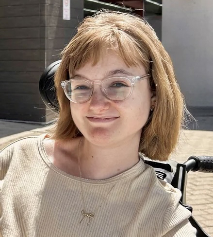 Hannah smiles, close-lipped, at the camera. She sits at a slight angle away from the camera. She is wearing a cream colored shirt. She has green eyes and strawberry blonde hair with bangs. Her hair hangs in a bob just above her shoulders. She has clear, round-framed glasses, double-pierced ears, and a gold necklace with a pendant in the shape of a bow. Behind her head, one can see the headrest of her wheelchair. She is sitting on brick pavers, and a brick building can be seen in the background.