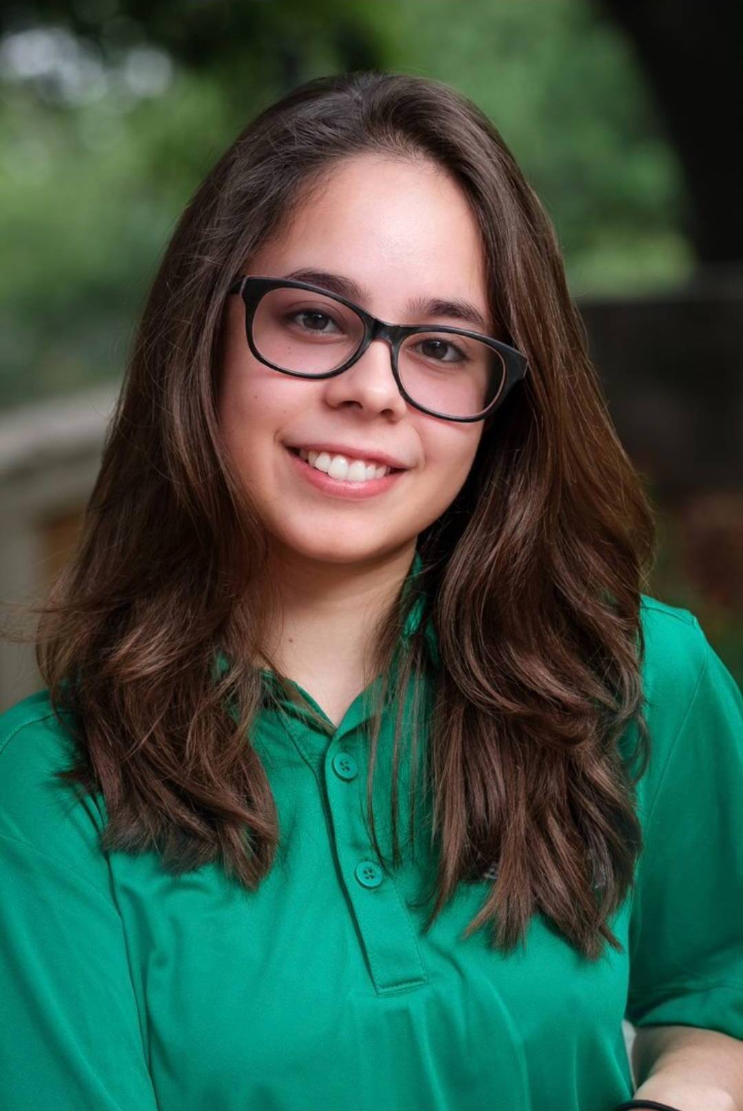Isabella smiles at the camera. She is wearing dark, square-framed glasses and a green polo shirt. She has long, wavy brown hair. In the background, which is blurred, one can see green foliage.