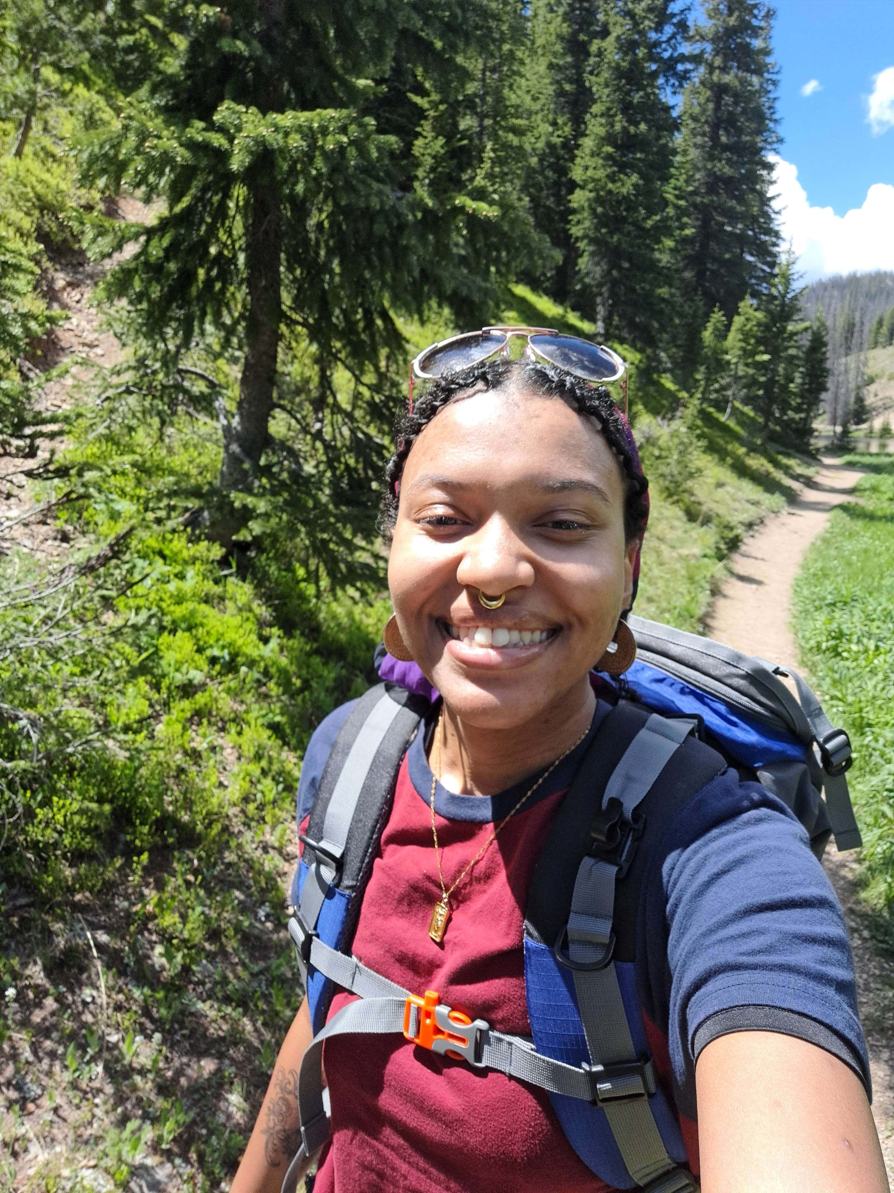 Jasmine smiles while taking a selfie. She has braided black hair pulled into a ponytail. She is hiking in a mountainous region. The pine trees, behind and to her right side and visible on the hillsides behind her, are a vibrant green. The dirt path on which she is walking extends behind her into the distance before disappearing behind the treeline. She has sunglasses on her head and is wearing a red and blue shirt as well as a blue and grey hiking bikepack with a chest strap. She has a septum piercing.