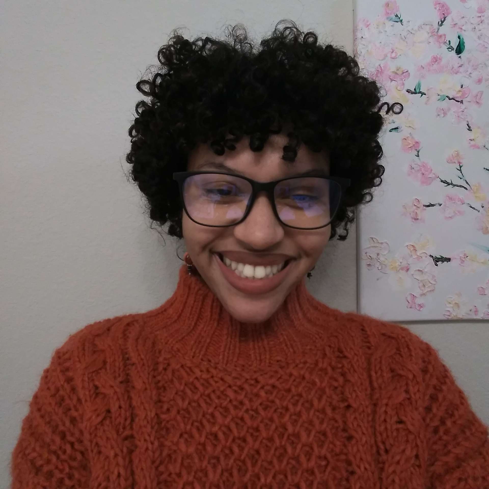 Jasmine smiles at the camera. She is standing in front of a white wall that has a floral painting on it, just visible over her left shoulder. She has black curly hair in a bob and is wearing dark, square-framed glasses and a red, cable-knit sweater.