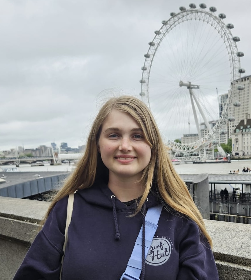 Kaitlyn stands, smiling at the camera. She has long, straight blonde hair and blue eyes. She is wearing a navy blue hooded sweatshirt and a light blue cross-body bag. Behind her, in the distance, is a large ferris wheel, a cityscape, and an overcast sky.