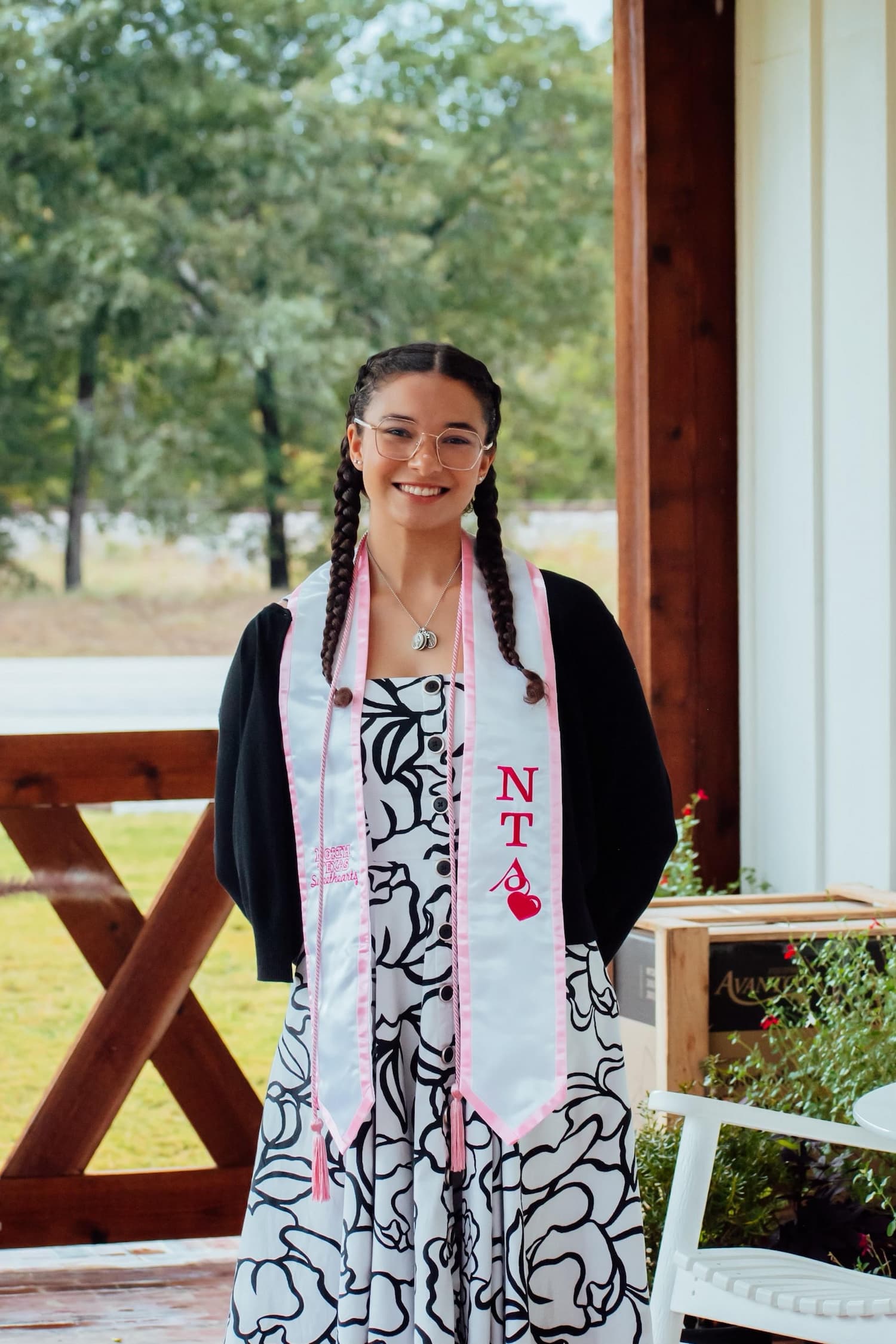 Karina is standing on a brick-paved patio next to a white patio chair, wearing a white dress with black stenciling and a black dress cardigan. She is wearing pink graduation chords and a white stole with a pink border and the acronym NTS. She has dark brown hair, braided into two pigtails. She is wearing a silver chain necklace with coin pendants. She is also wearing square, thin-framed glasses. In the background, one can see a wooden gate and trees..