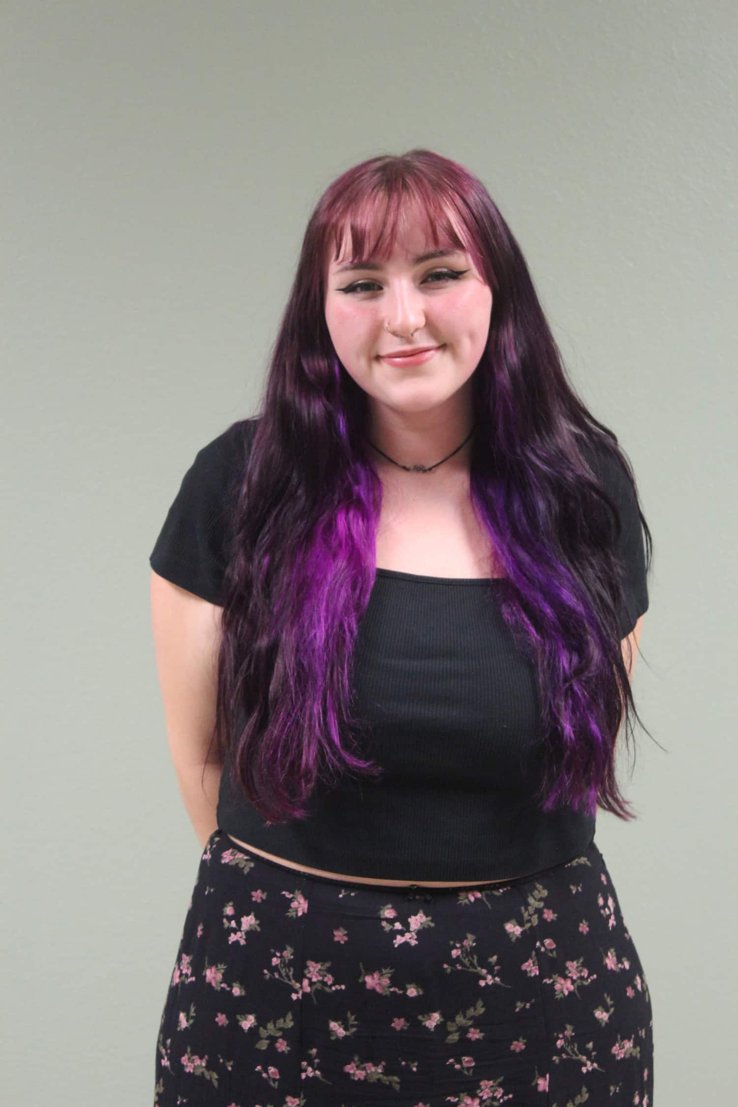 Kyhlie stands, arms behind her back, and smiles at the camera. She is wearing a black top and a black and pink floral skirt. Her hair is long and straight, and she has cut-across bangs. Her hair is dyed, with a rose pink on top and deep purple in the bottom layers. The background behind her is a white wall.