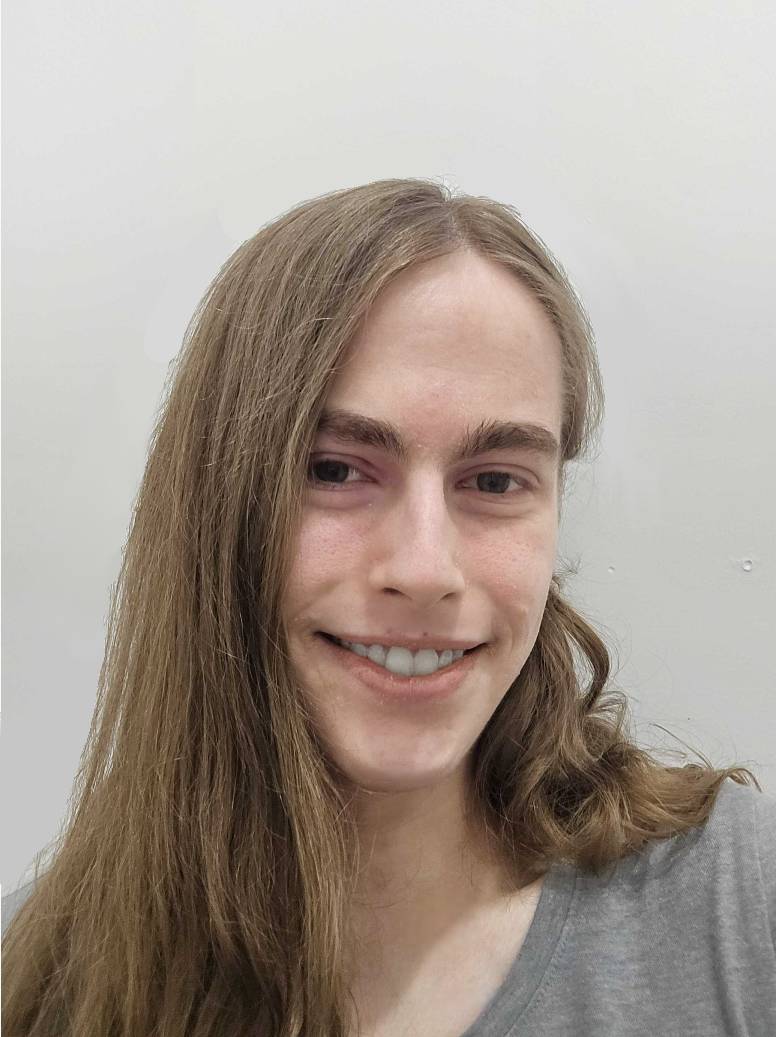 Lily smiles at the camera. She has long, lighter brown hair and is wearing a grey shirt.