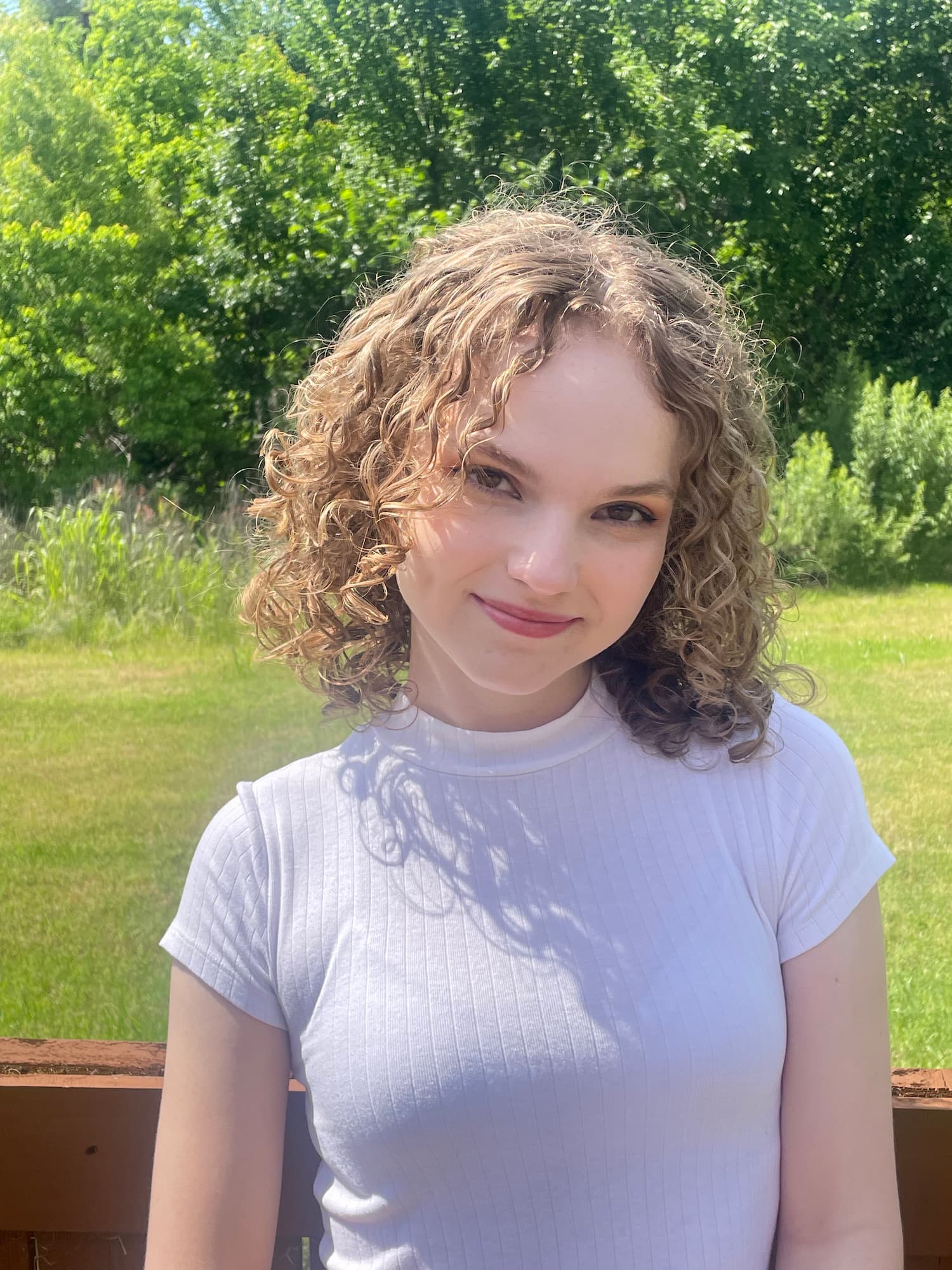 Mallory stands in front of a wooden fence; behind it is a green field with brush and trees in the distance. She is wearing a white shirt with a high neckline. She has curly blonde hair that reaches just to her shoulders. She has fair skin and dark brown eyes. She is smiling (close-lipped) at the camera with her head slightly cocked to the side.