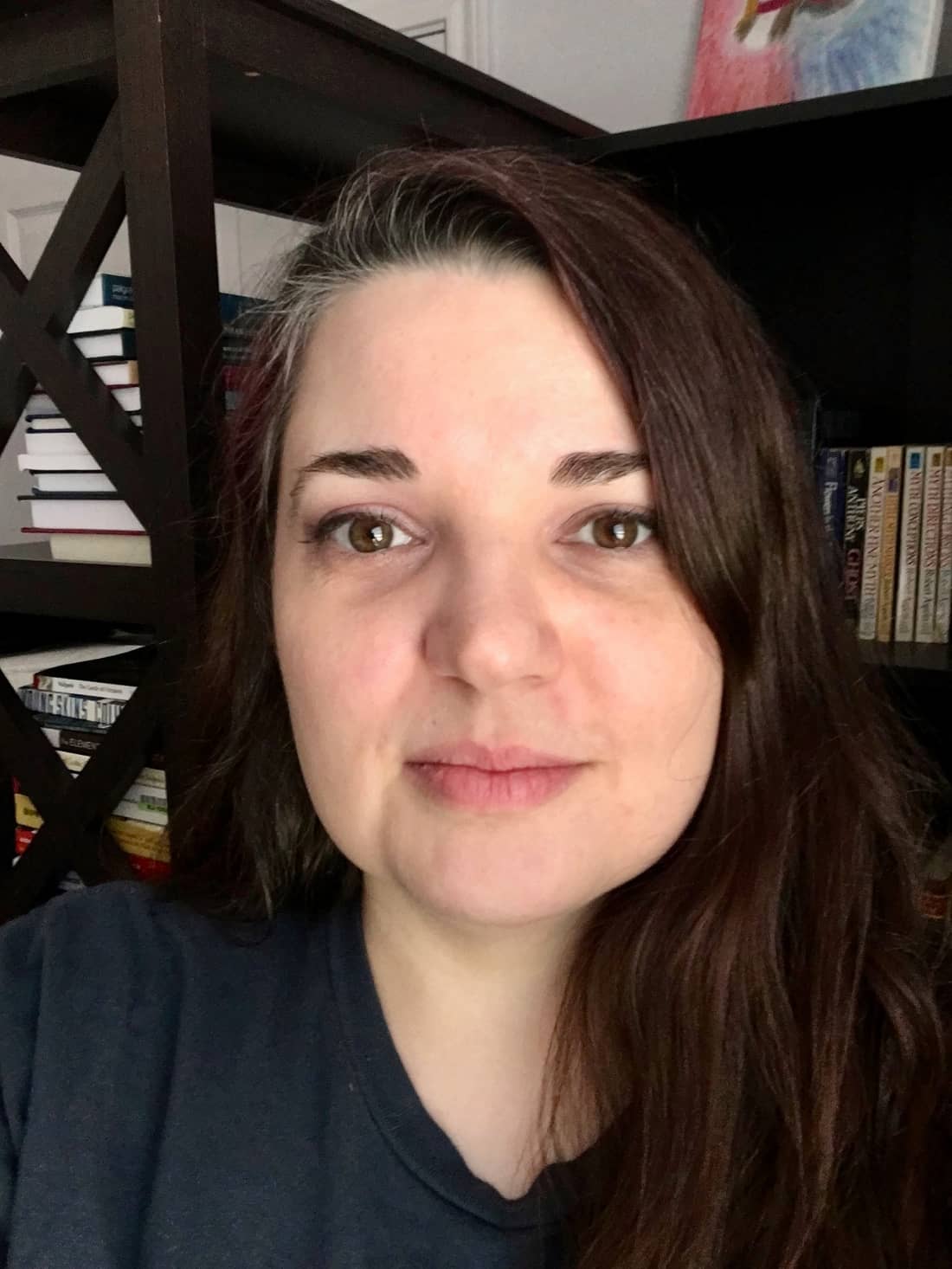 Mandy smiles, close-lipped, at the camera. She has dark brown, straight hair and dark brown eyes. She is wearing a black shirt and is sitting in front of two dark wood bookshelves.