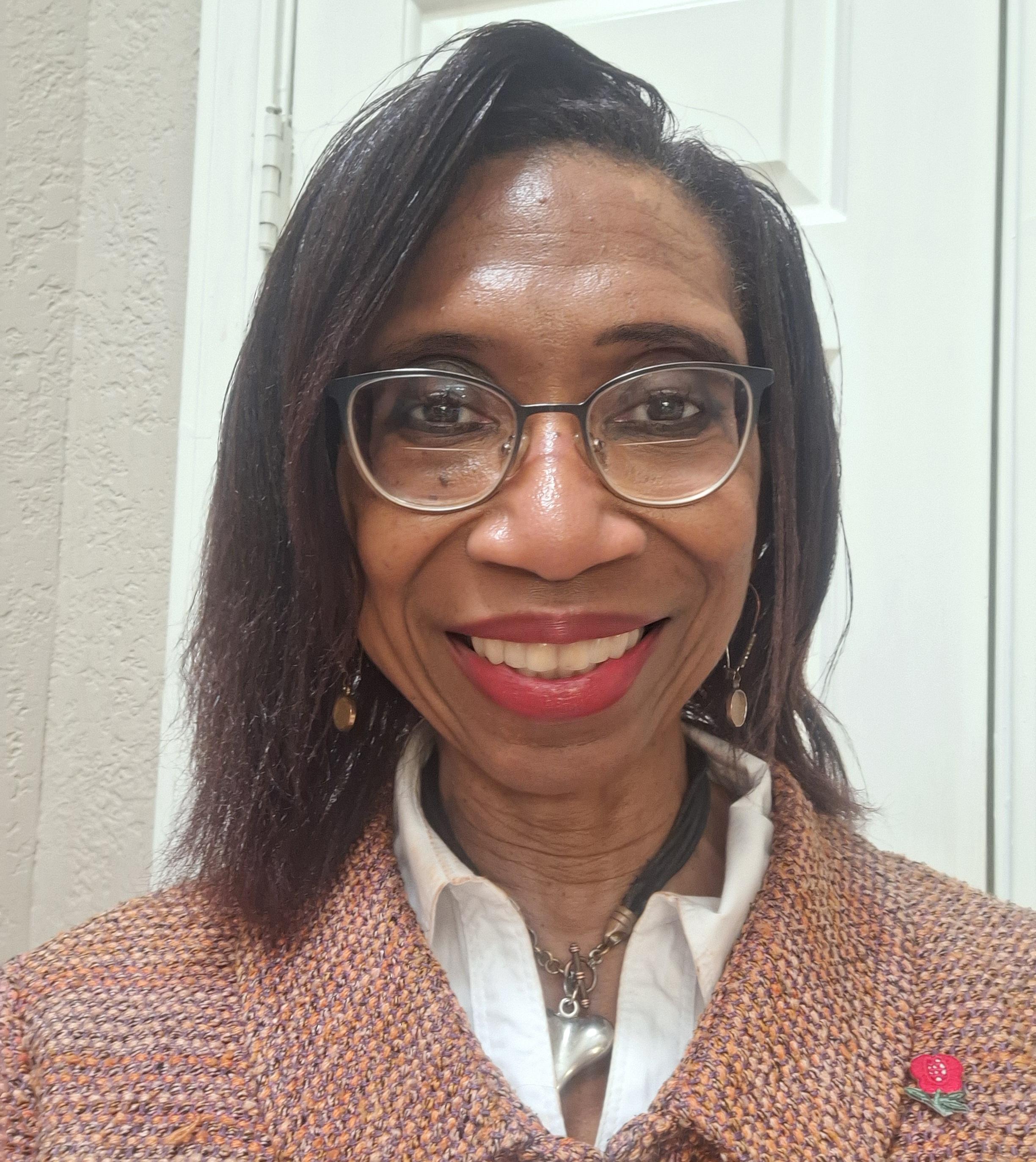 Regina stands in front of a white door, smiling widely at the camera. She is wearing circular glasses with black frames, dangling eaarings, and a necklace with a silver pendant. Her dark brown hair is straightened and falls just to her shoulders. She is wearing a white collared shirt and a pink and salmon-colored, knitted blazer. Pinned on the blazer is a metal pin in the shape of a pink flower.