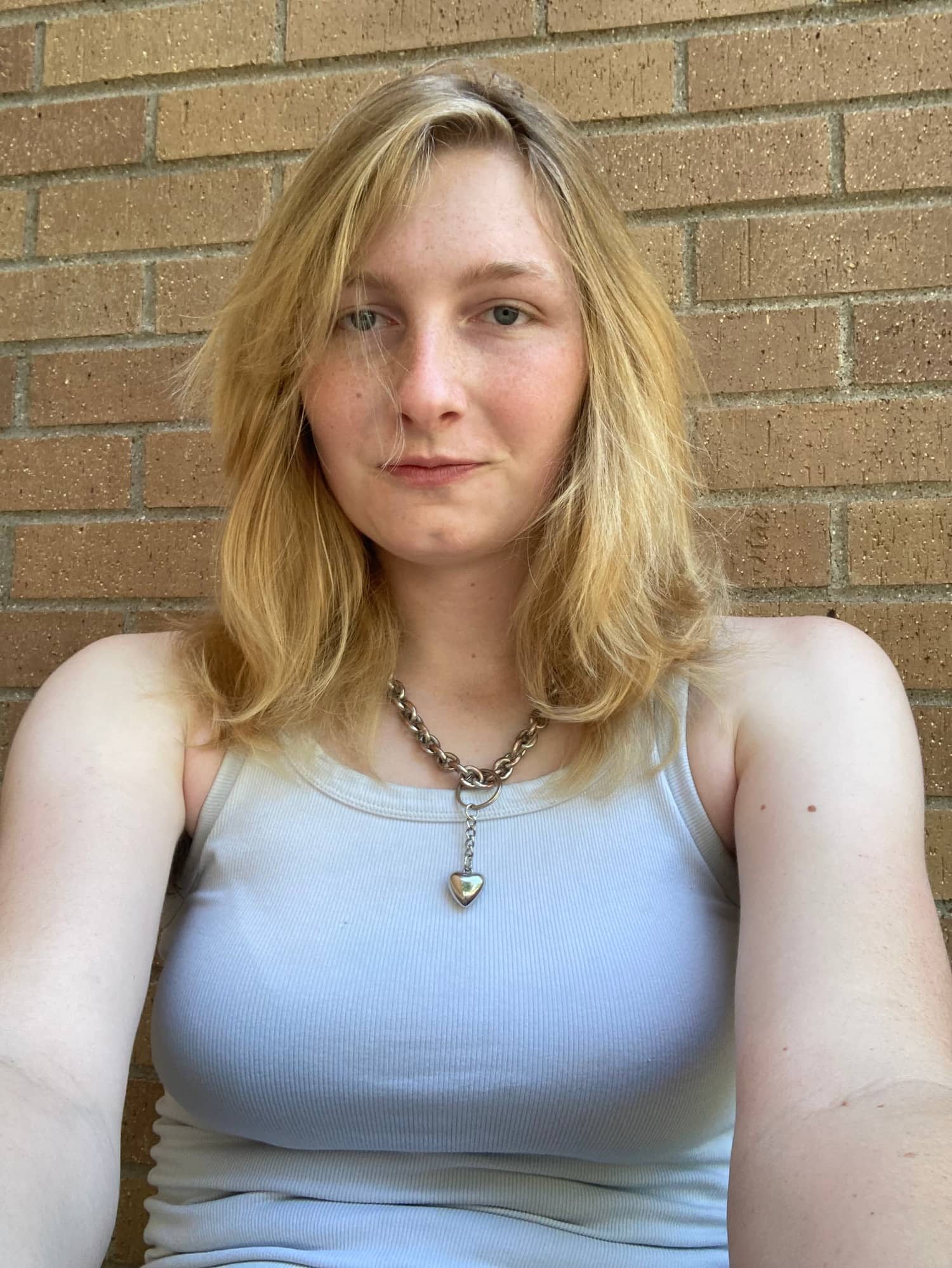 Rose looks at the camera. She is standing in front of a brick wall. She has medium-length, blond hair and blue eyes. She is wearing a white tank top and a silver chain necklace with a heart pendant.