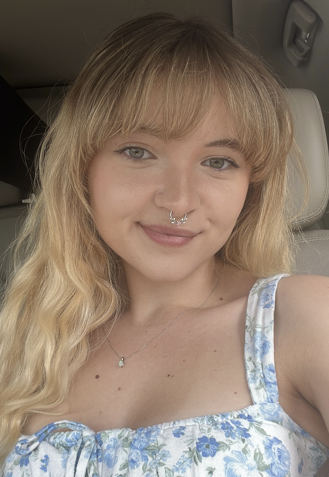 Ryleigh smiles (close-lipped) at the camera. She has long wavy blonde hair with bangs that are cut across her forehead. She had green eyes. She is wearing a white dress shirt with a floral pattern and a dainty silver necklace with a small opal pendant. She has a septum piercing in her nose, the jewelry itself is silver with leaf embellishments. In the background, one can see she is sitting in her car.