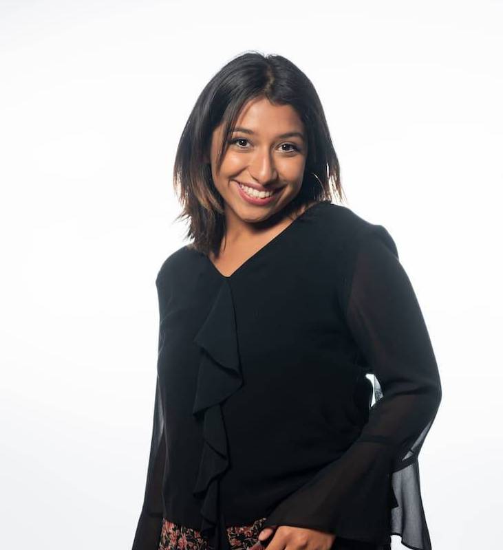 Sara stands and smiles at the camera. She has black hair and dark brown eyes. She is wearing a black dress shirt with long, sheer sleeves. Her skirt is floral with black, red, beige, and white tones. In the background is a white screen.