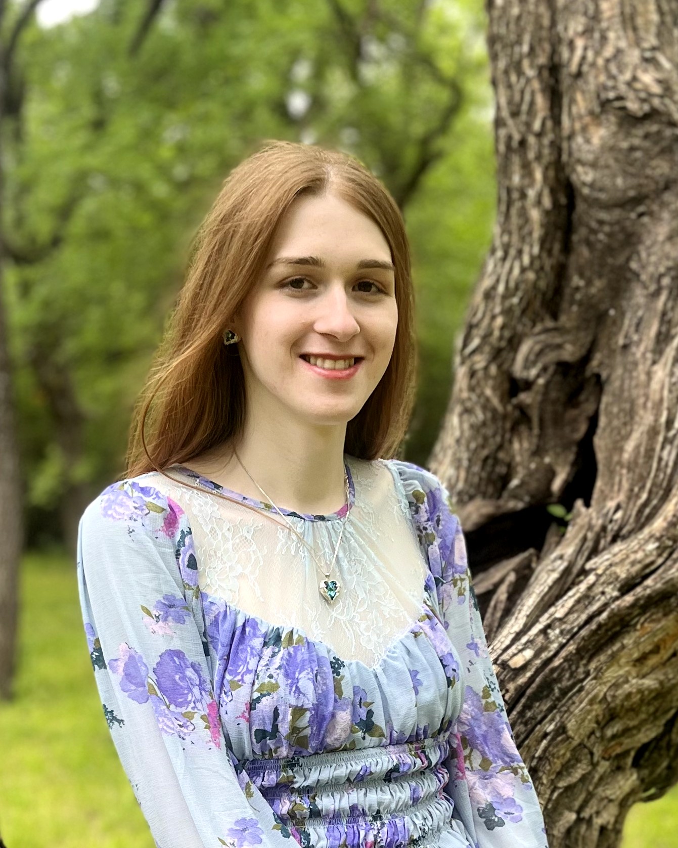 Sora stands, slightly turned from the camera, in front of an old tree. She is looking at the camera and smiling. Her long, red hair is hanging straight behind her back.She is wearing a periwinkle dress with floral embellishments and a sheer lace cut-out near the neck. She is also wearing a silver heart pendant necklace with a green jewel and matching heart earrings. In the blurred background, one can see more green trees.