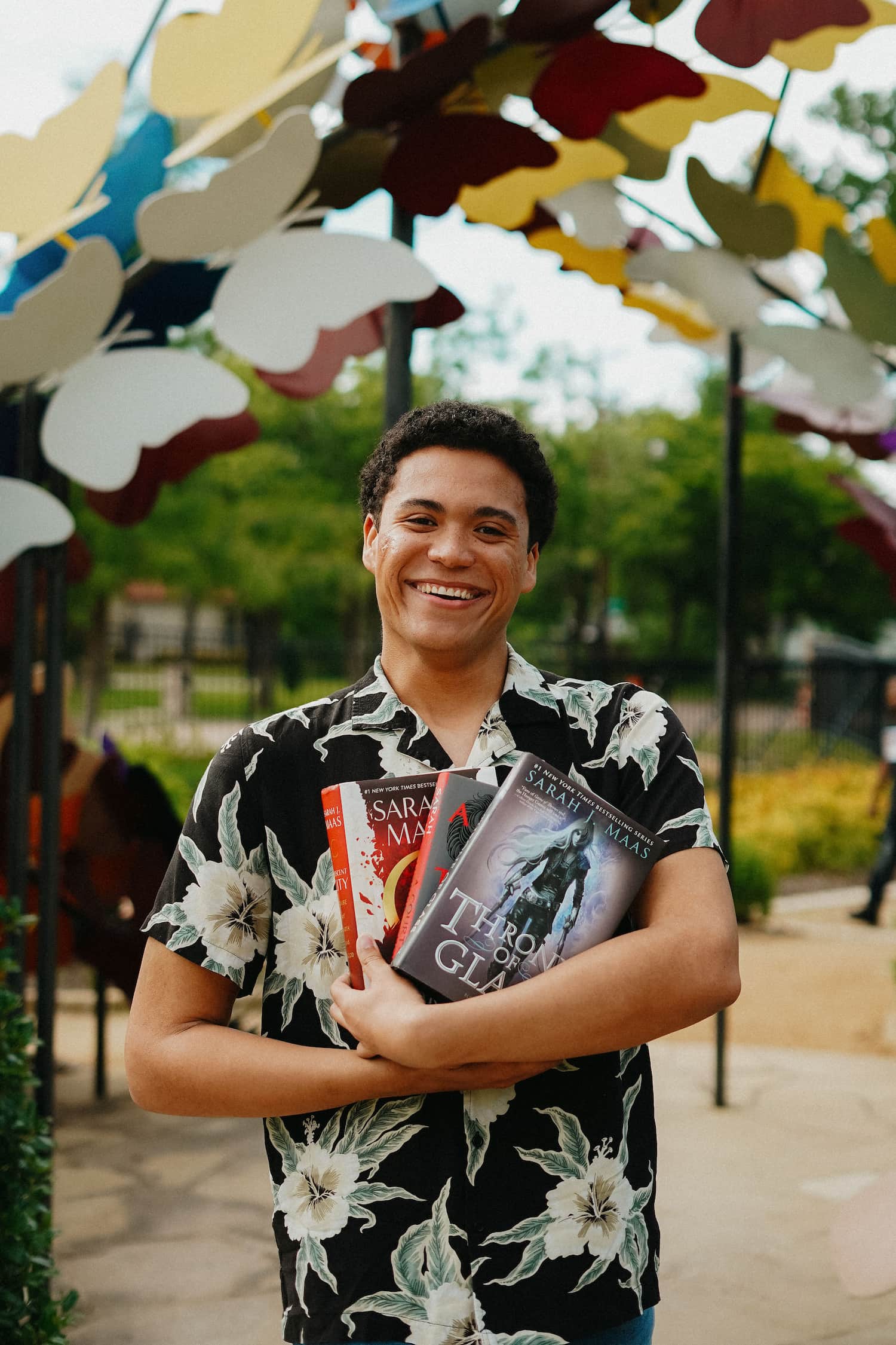 Thomasin stands, looking at the camera. He is smiling widely and has dark brown curly hair and dark brown eyes. He is holding three books by Sarah J. Maas and is wearing a black button-up shirt with white florals. In the blurred background, one can see a butterfly arch above him and a green garden with trees.