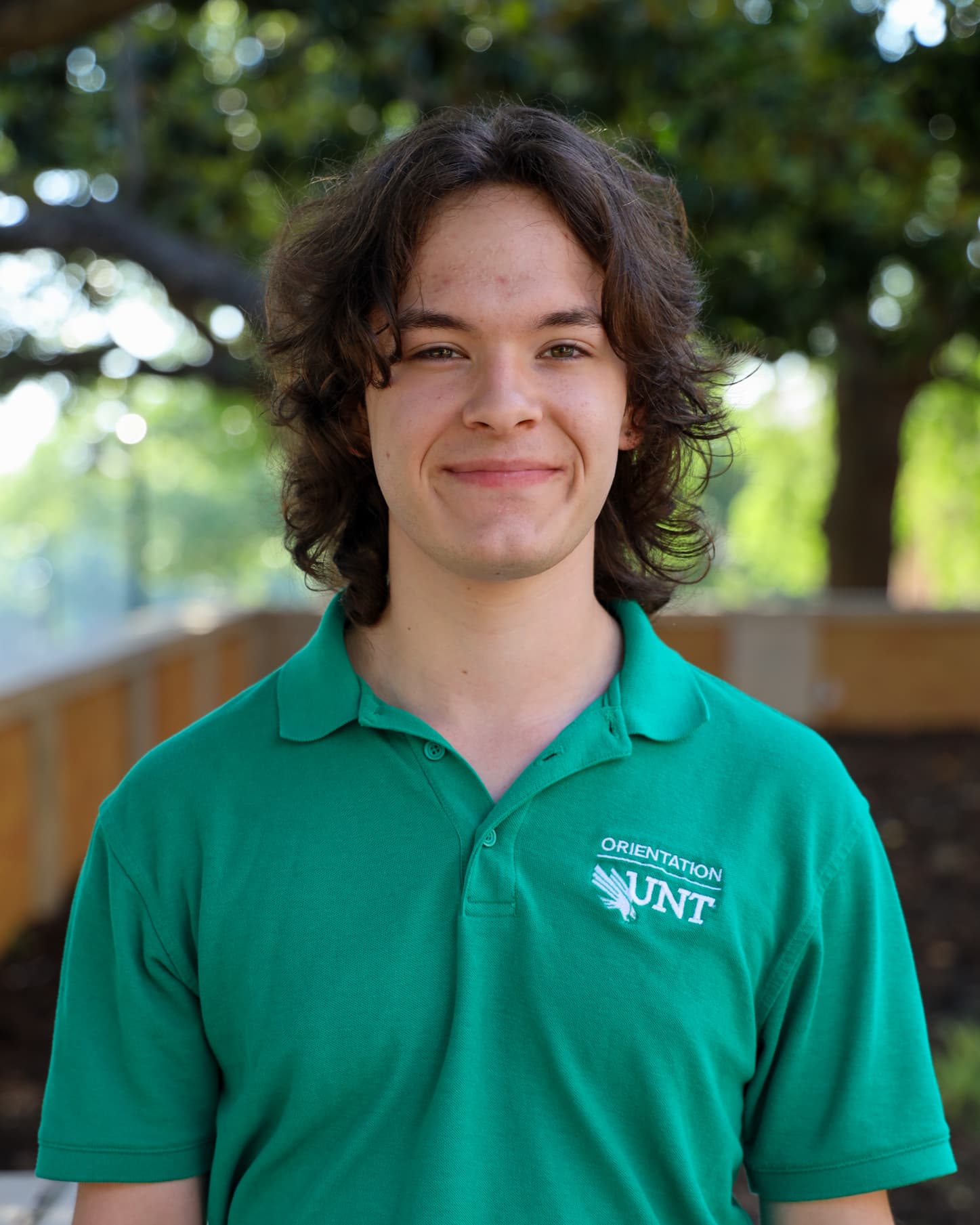 Trey is smiling, close-lipped, at the camera. He is wearing a UNT Orientation, green polo shirt. He has dark brown, wavy, layered hair that falls just above his shoulders. In the background, which is blurred, one can see a wooden fence and trees.